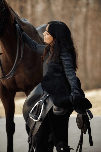 Woman wearing Bella pull on riding leggings turned toward her horse smiling while holding her dressage saddle. 