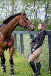 Woman standing with her horse outside while wearing the GG turtleneck with built in earbud pockets and cord keepers, as well as dun colored May Babes innovative breeches with built in uderwear. 