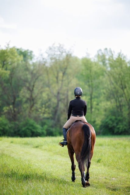 Model in a black turrleneck wearing the Off course breeches in Dun rides her bay horse away from the camera with a green scenery in the background.