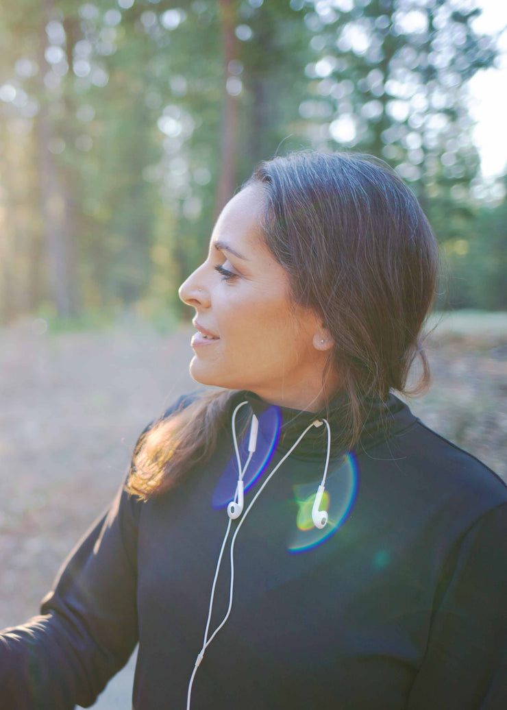 Woman of Indian descent wears black GG technical shirt in black. She looks off into the sunset while wearing her corded earbuds through the keeps on the neck of the shirt with them hanging down to the side.  