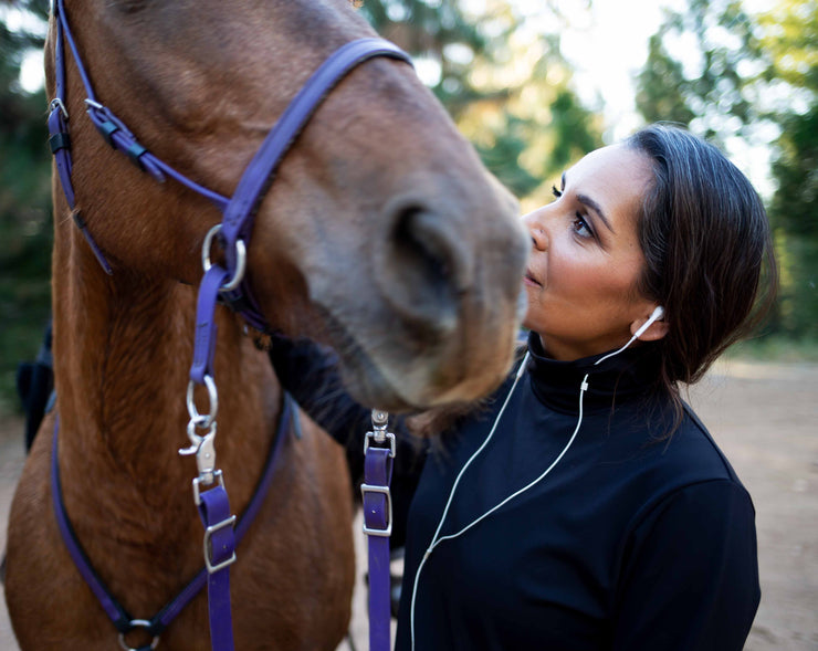 Woman of Indian descent wears a black GG Technical shirt in black, with corded earbuds running through keepers and into her ears. She gazes up to her horse who is standing to her right, wearing a purple headstall, reins and breast collar. 