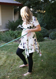 Pregnant woman wearing black maternity leggings and patterned floral knee length dress. She is pulling a hose while walking backward on her lawn. She is wearing size small autumn maternity leggings. 