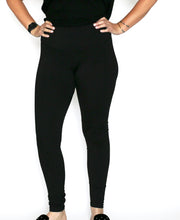 Woman in front of white seamless background standing toward the camera, wearing black equestrian workout leggings with slide shoes. Her hands are on her hips.