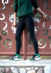 Woman in black May Babes equestrian workout leggings with built in under garment, facing the camera. She is wearing green Nike exercise shoes and a matching green sweat shirt. She is standing in front of a laser cut metal fence, with a concrete and stone base.