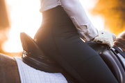 Woman equestrian sitting in a dressage saddle wearing May Babes riding breeches in black. Her saddle is black and she is wearing white equestrian riding gloves and a white equestrian show blouse. The sunset is behind them.