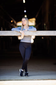 Woman wearing black Phoenix legggings with her legs crossed while standing and learning against a board inside a barn. She is wearing a grey long sleeve shirt that highlights the high waist of the leggings, and holding a coffee in her right hand while looking off into the distance. 