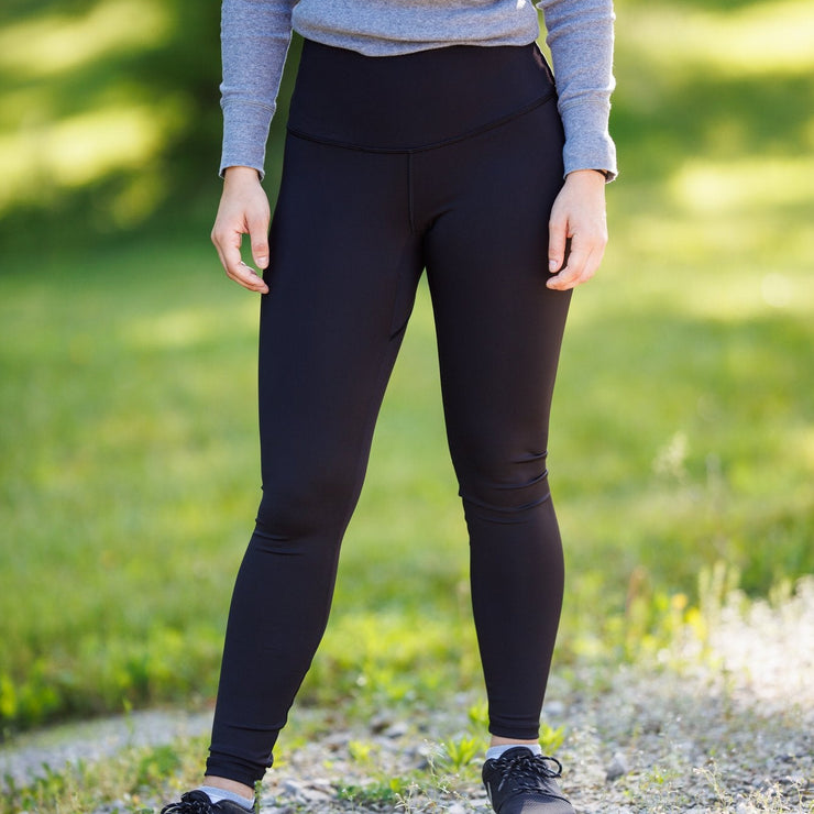 Woman in black Phoenix leggings standing to the side of a road near green grass. She is wearing a grey long sleeved shirt that shows the high waist of the leggings. You only see from chest level down.
