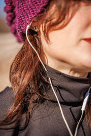Woman in black GG Technical Shirt wears her earbuds in her ears threaded through the cord keeper of the turtleneck. She wears a purple beanie and you only see a portion of her face. 