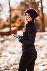 Woman in black GG Technical shirt turtleneck and black Phoenix leggings stands in the snow while holding her phone, and turned toward the camera. She is wearing a purple beanie and has earbuds in her ears.