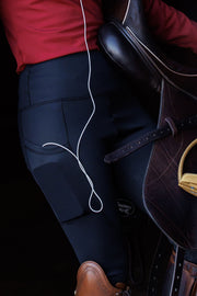 Woman in a red GG Technical Shirt and Ges breeches leans against a wall while holding a dressage saddle. She has a phone in her right pocket with earbuds and cords leading upward. 