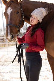 Woman in a red GG Technical shirt and black May Babes leggings stands with her horse in the snow. She holds a cup of coffee and is wearing a tan knitted beanie while smiling at her horse