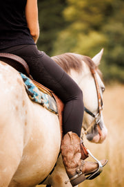 Woman in a western saddle on top of an apaloosa horse. She is wearing black May Babes riding leggings with brown western pull on boots and spurs. You see mostly just her lower half with her horse in the distance.