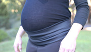 Close up detail image of a pregnant woman's waist area. Pregnant woman wearing black Autumn maternity leggings. The waistband is folded down underneath her bump, with a black casual blouse tucked into the leggings. 
